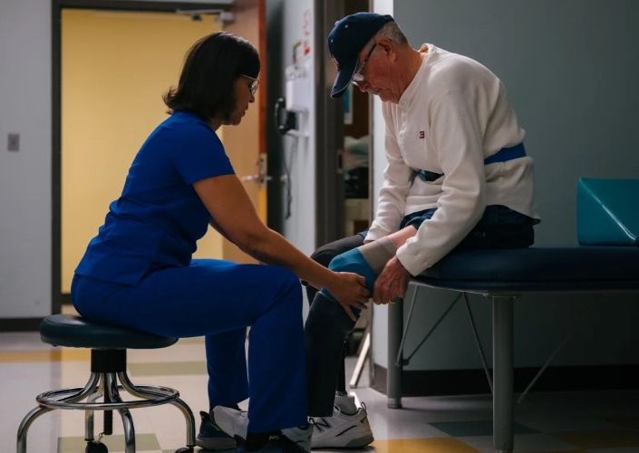 A nurse in blue scrubs helps rehab an elderly man's knee at Siskin Hospital for Physical Rehabilitation where they use SONIFI Health digital whiteboards for care coordination and communication
